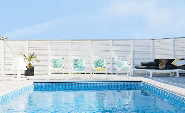 En pool med blått kakel omgärdat av ett trädäck med vita möbler och ett högt vitt staket.