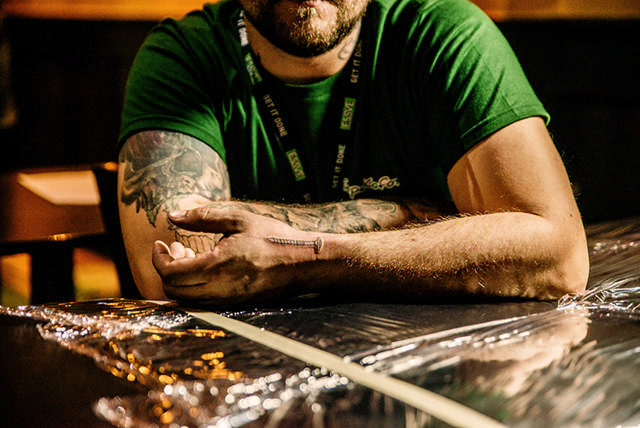 Vīrietis sēž pie tetovēšanas galda un rāda savu jauno tetovējumu ar celtniecības skrūvi uz rokas.